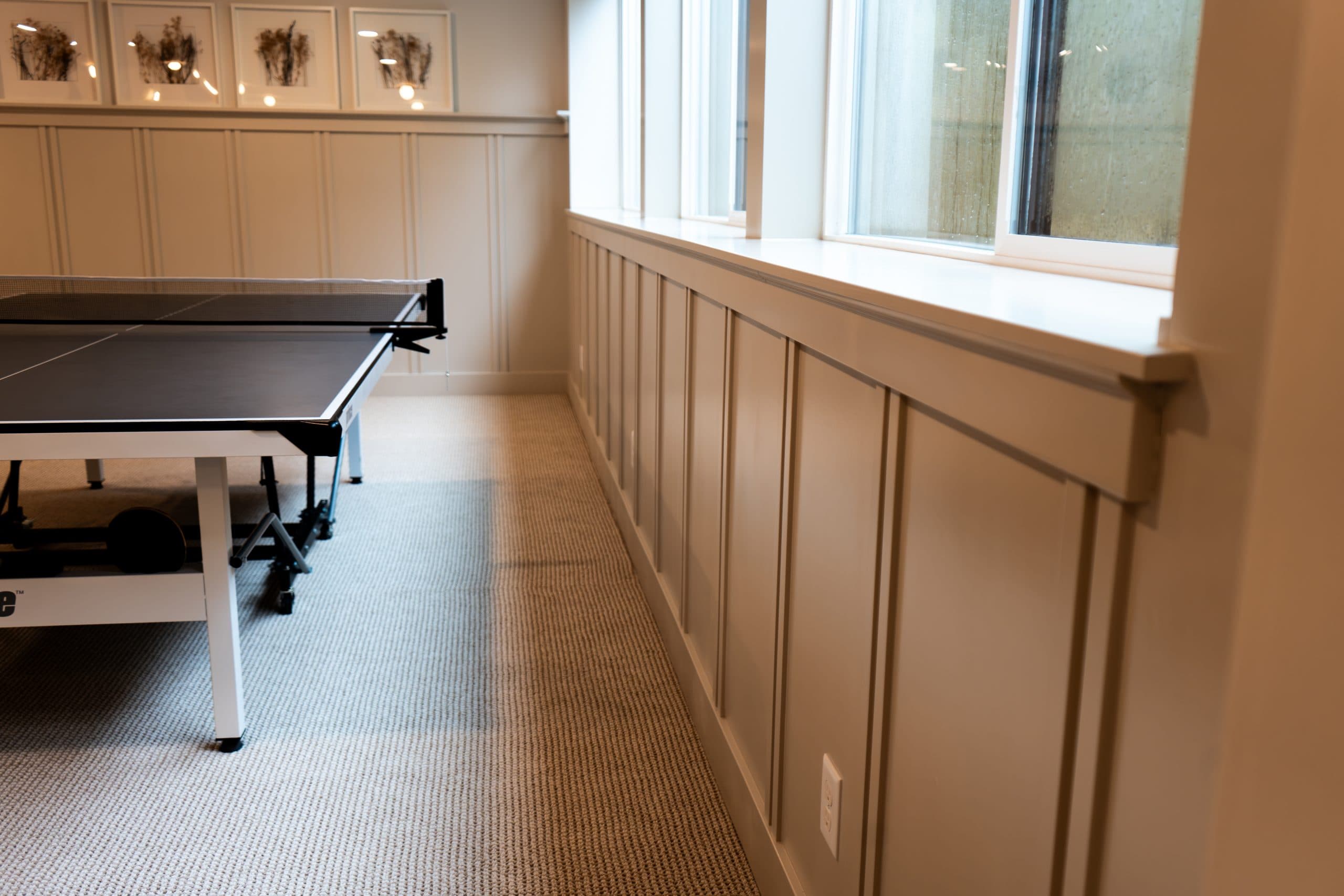 A ping pong table is positioned in a well-lit room with large windows, carpeted flooring, decorative wainscoting, and elegant interior painting—reflecting the charm of residential painting styles found in Ada Michigan.