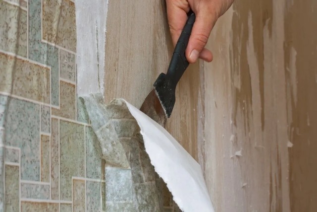 A hand uses a scraper to peel wallpaper off a wall in a Caledonia, Michigan home, revealing a patterned green and beige layer underneath—perfect prep for quality interior painting.