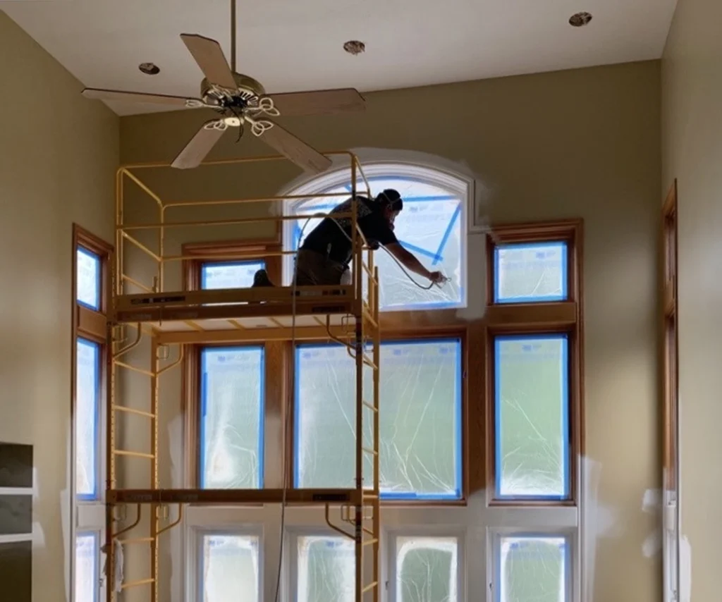 A person stands on scaffolding, taping plastic sheets over tall windows in a room with high ceilings and a ceiling fan—prepping for a residential painting project in Ada, Michigan.