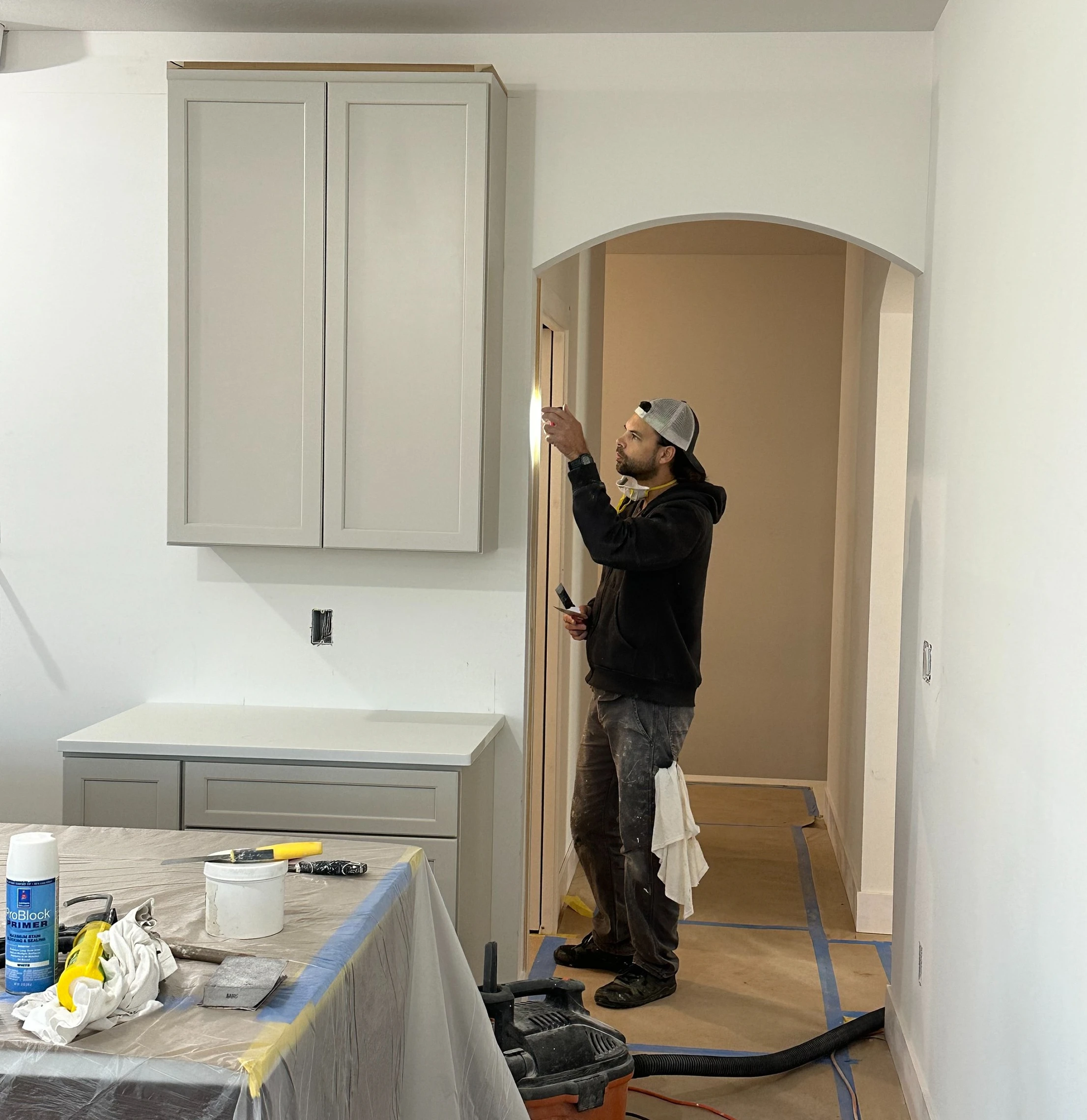 A man in work clothes stands under an archway, inspecting a doorway in a room under renovation, where tools and cabinets suggest ongoing interior painting and cabinet painting work.