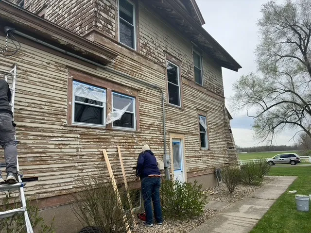 Two people work on the exterior of an old house with peeling paint and exposed wood, surrounded by shrubs and a cloudy sky, showcasing residential painting skills often seen in Ada Michigan.