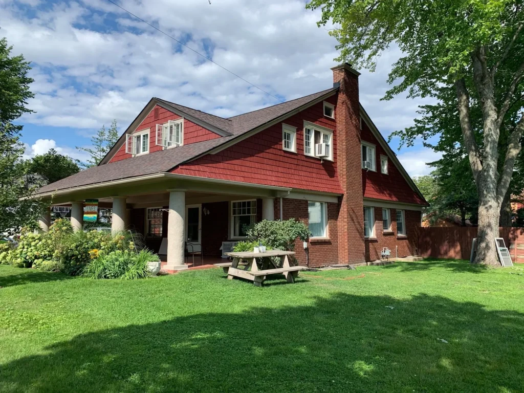 A red brick and wood house with a large porch, white columns, dormer windows, and a picnic table on a green lawn under a partly cloudy sky—perfect for residential painting projects in Ada Michigan.