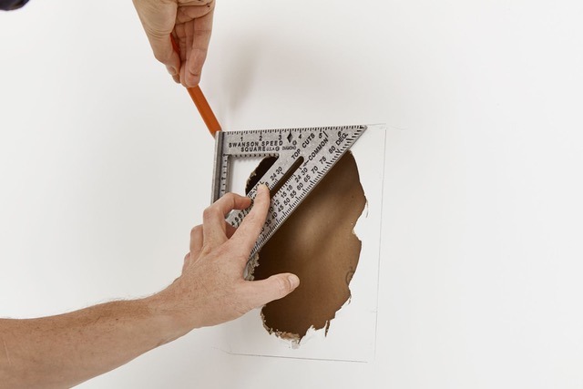 A person uses a carpenter's square and pencil to mark a rectangular outline around a hole in a white wall, preparing the area for residential painting.