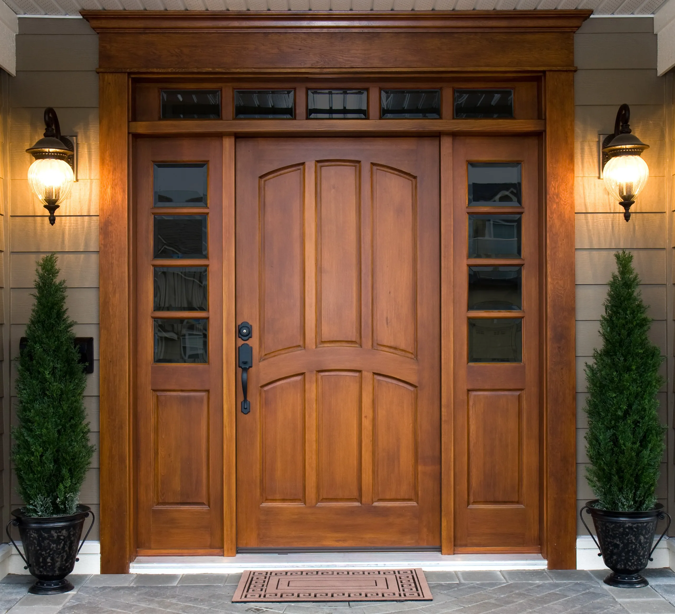 A wooden front door with glass panels, two side windows, two outdoor wall lanterns, and two potted evergreen plants on either side—perfectly complemented by expert exterior painting for a welcoming residential entryway.