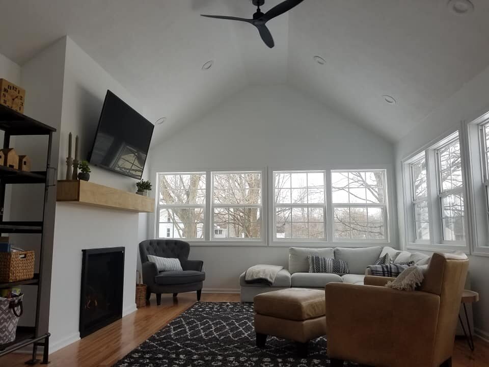 A modern living room with large windows, a beige sectional sofa, an armchair, a fireplace with a mantel, wall-mounted TV, and a black ceiling fan. - Jeff Bryant Painting