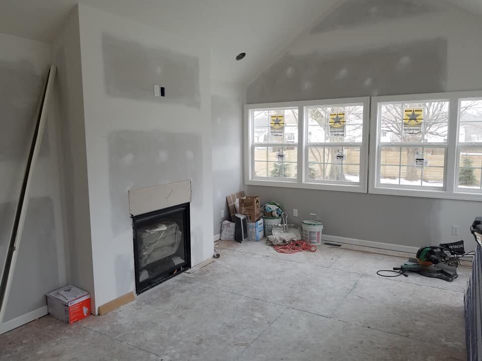 A room under construction with unfinished drywall, a fireplace, tools, boxes, and supplies scattered near large windows. - Jeff Bryant Painting