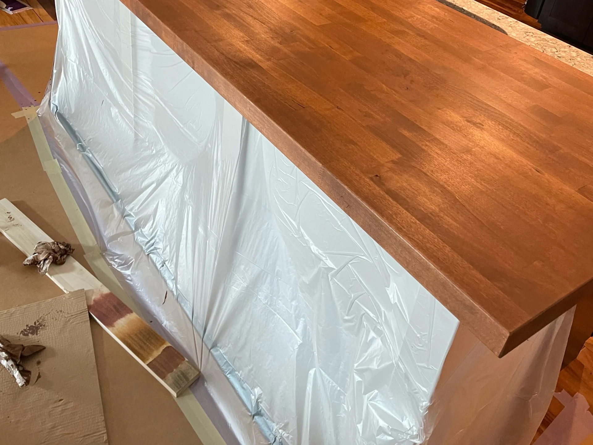 A wooden countertop with a stained finish is shown atop a base covered in protective plastic sheeting, with painting supplies and samples nearby. - Jeff Bryant Painting