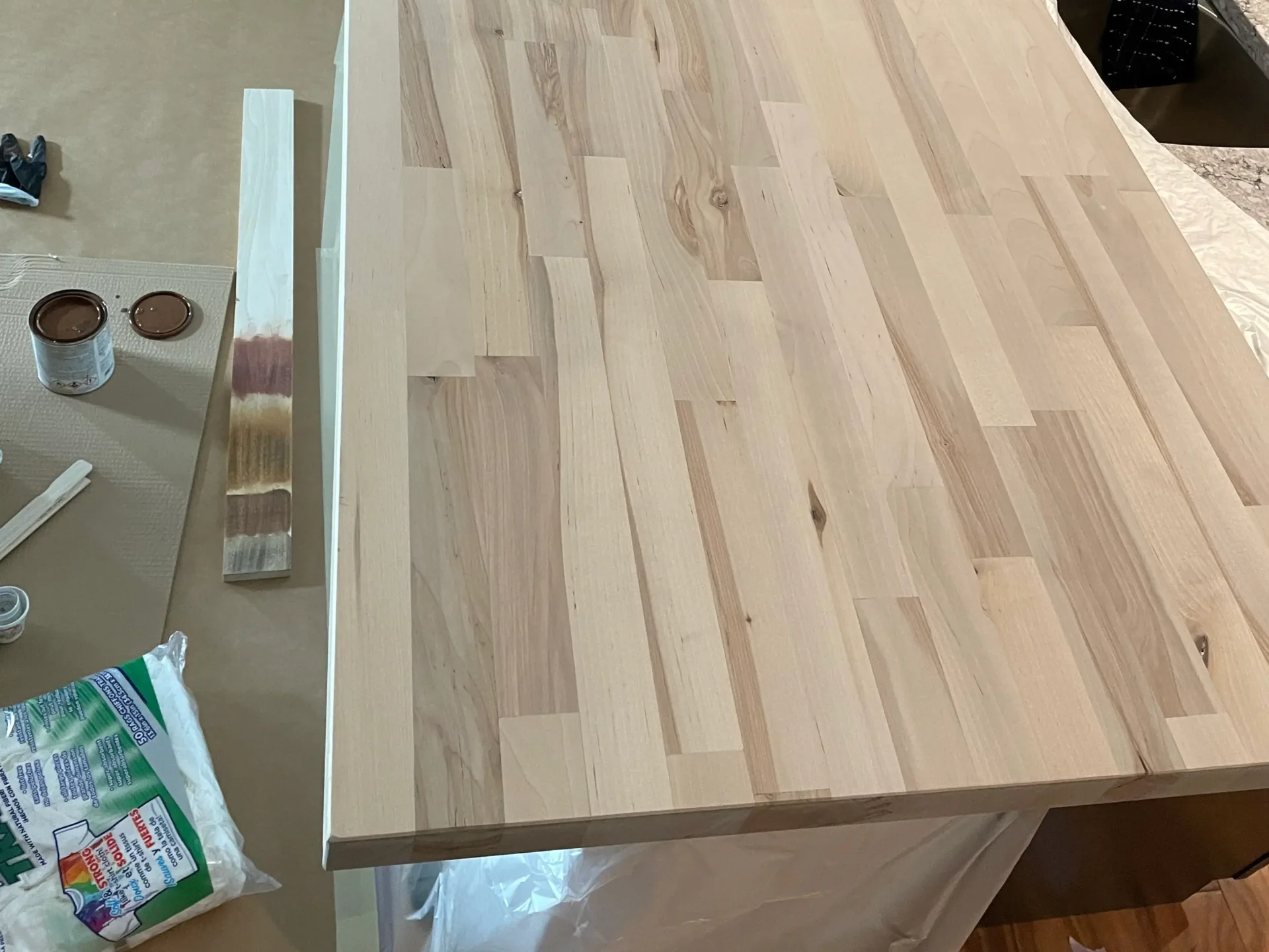 A light-colored, unfinished wood countertop sits on a work surface with wood stain cans, a test strip, and gloves nearby, prepared for staining. - Jeff Bryant Painting