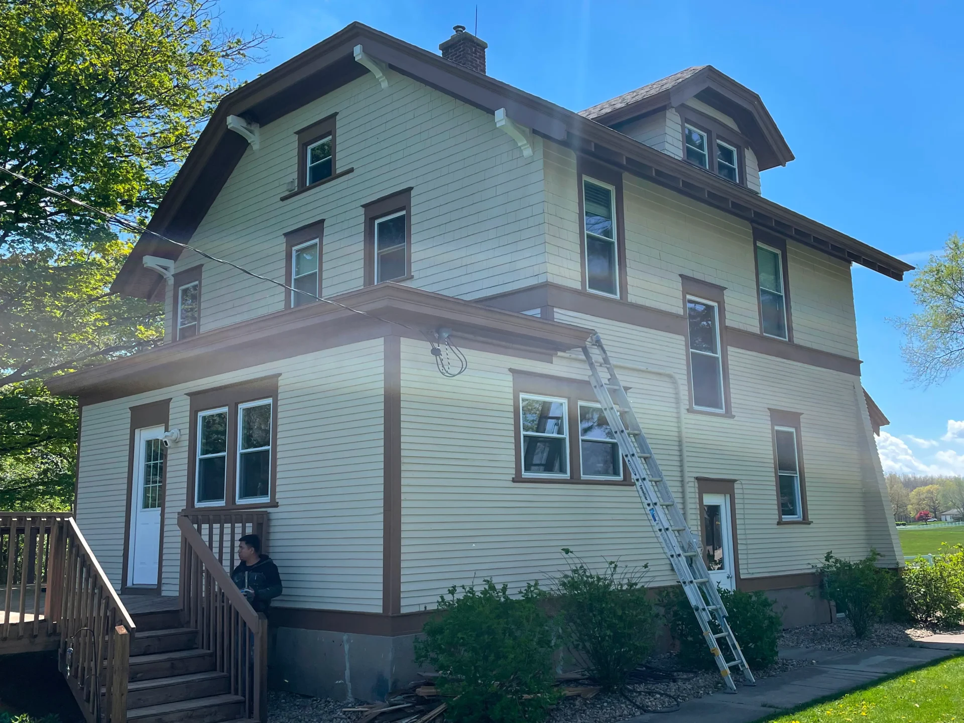 A beige two-story house with brown trim, a ladder leaning against the wall, and a person sitting by the deck stairway on a sunny day. - Jeff Bryant Painting