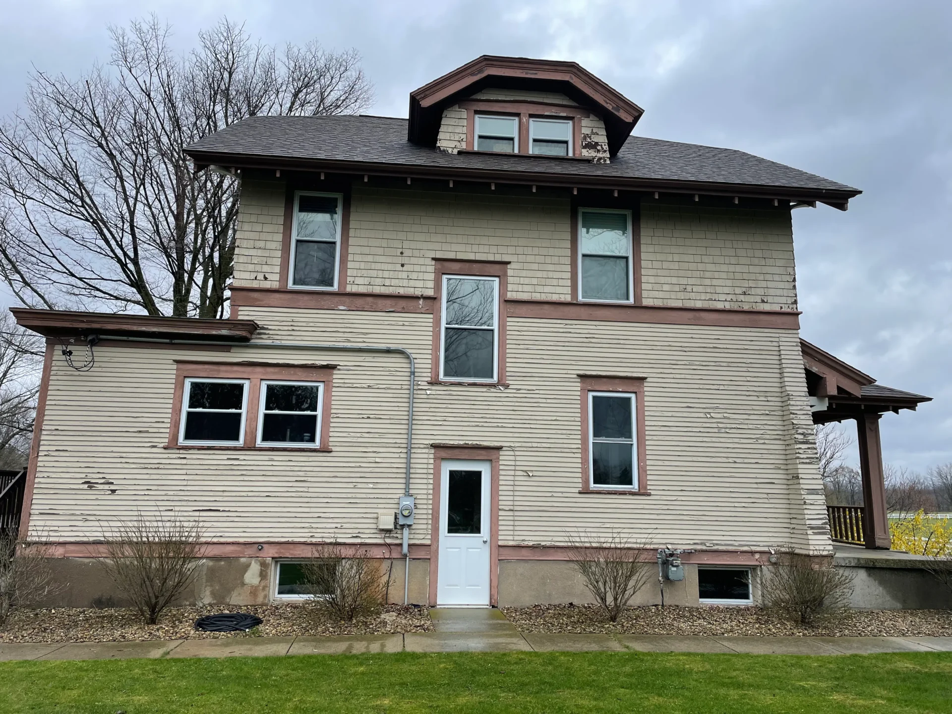 A two-story house with weathered beige siding, brown trim, and a small covered porch on a cloudy day. - Jeff Bryant Painting