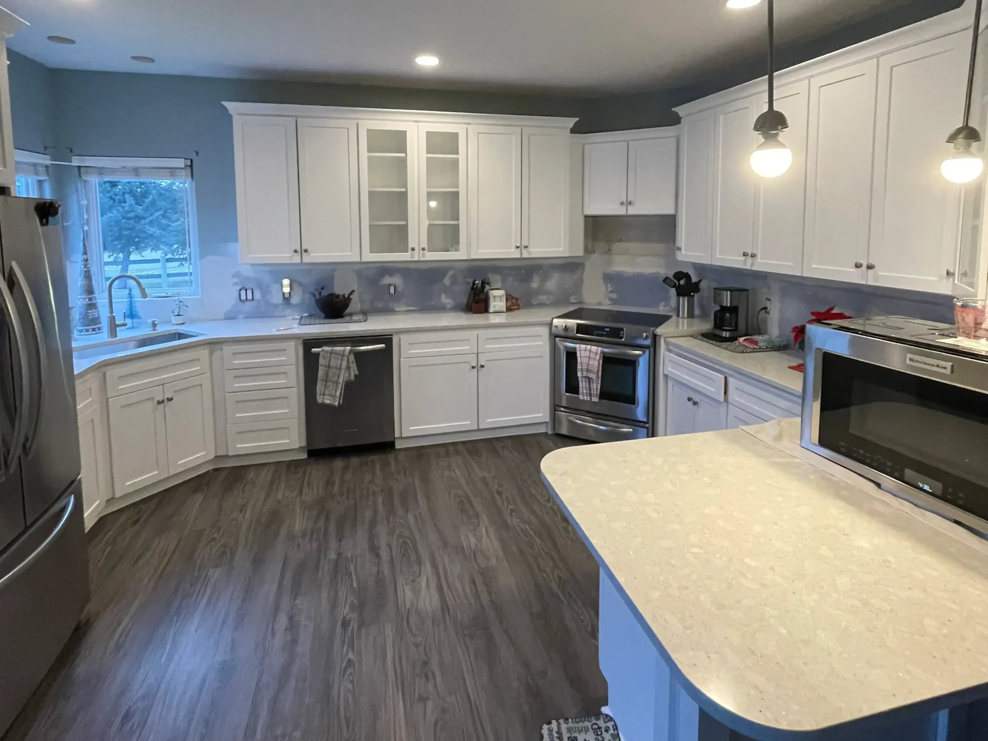 Modern kitchen with white cabinets, stainless steel appliances, light countertops, pendant lights, and wood-style flooring. A window is above the sink and various kitchen items are on the counters. - Jeff Bryant Painting