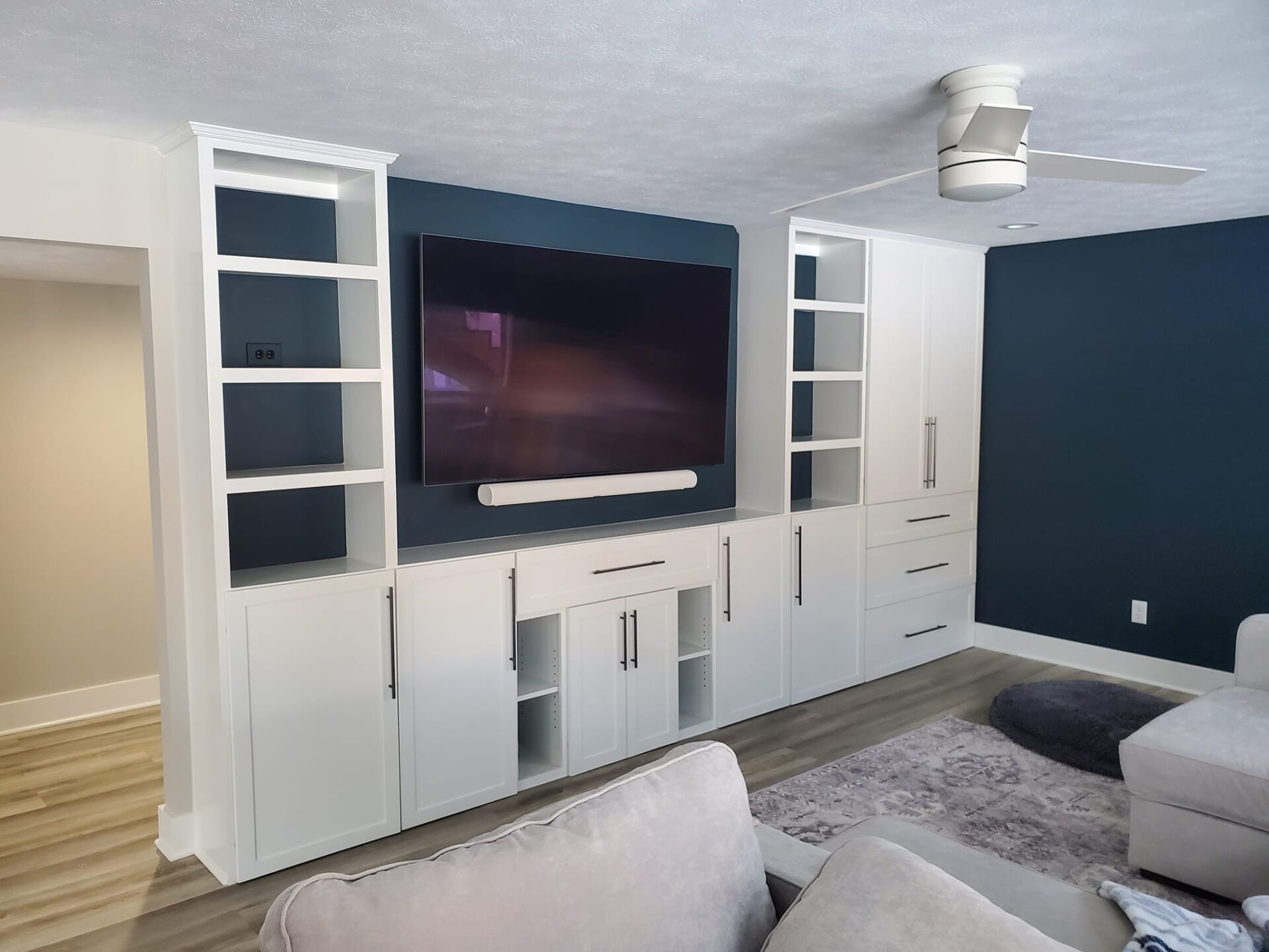 Modern living room with built-in white cabinets and shelves, a mounted flat-screen TV, a soundbar, and a ceiling fan against dark blue accent walls. - Jeff Bryant Painting