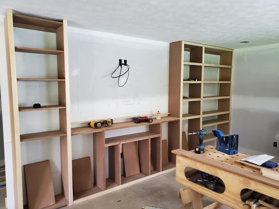 Unfinished built-in wooden shelves and cabinets under construction, with tools and materials on a workbench and shelf surfaces. - Jeff Bryant Painting