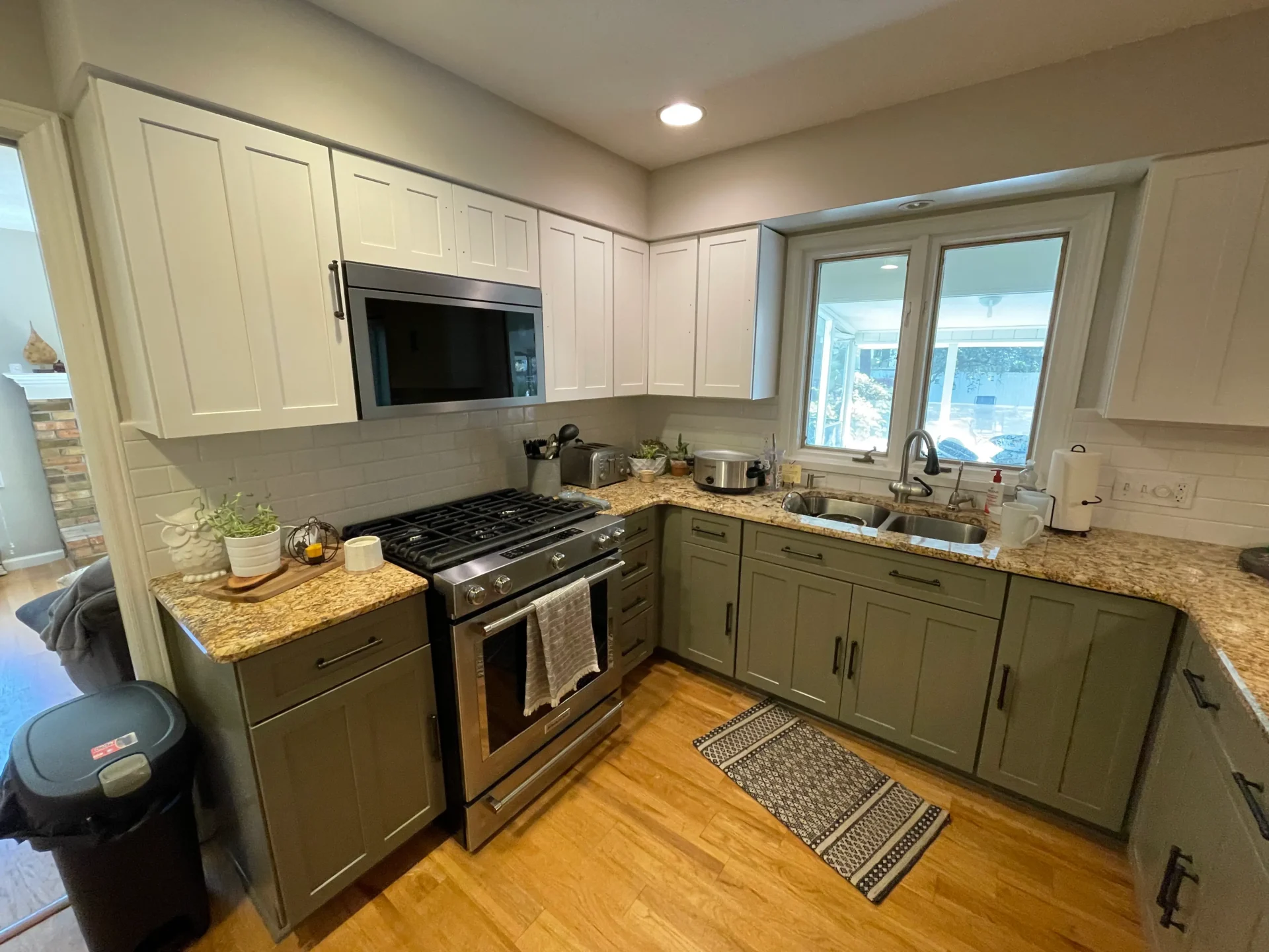Modern kitchen with green lower cabinets, white upper cabinets, granite countertops, stainless steel appliances, and a window above the sink. Various kitchen items are on the counters. - Jeff Bryant Painting