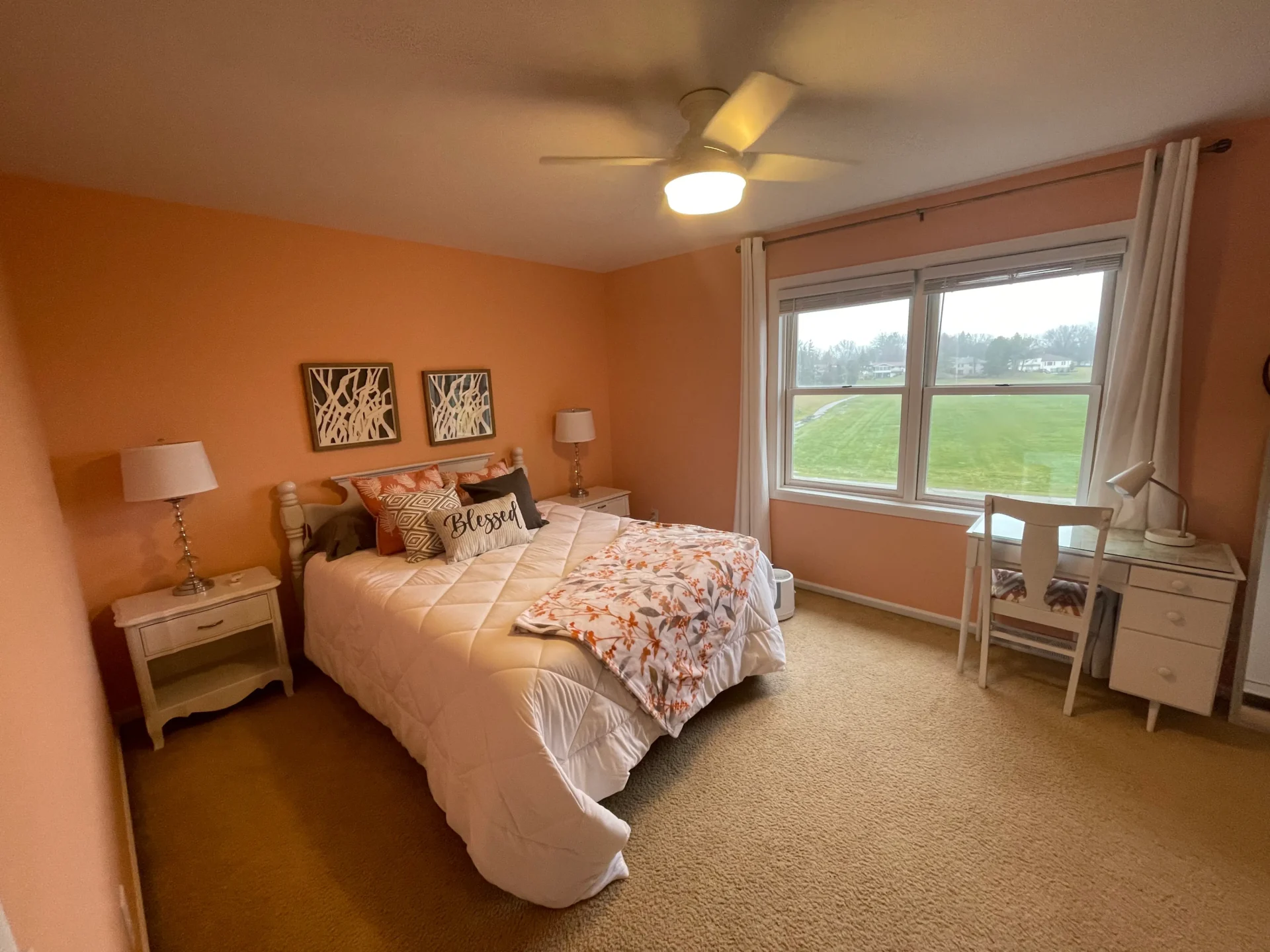 A neatly made bed with white and orange bedding in a peach-colored bedroom with two nightstands, a desk by the window, and a ceiling fan overhead. - Jeff Bryant Painting