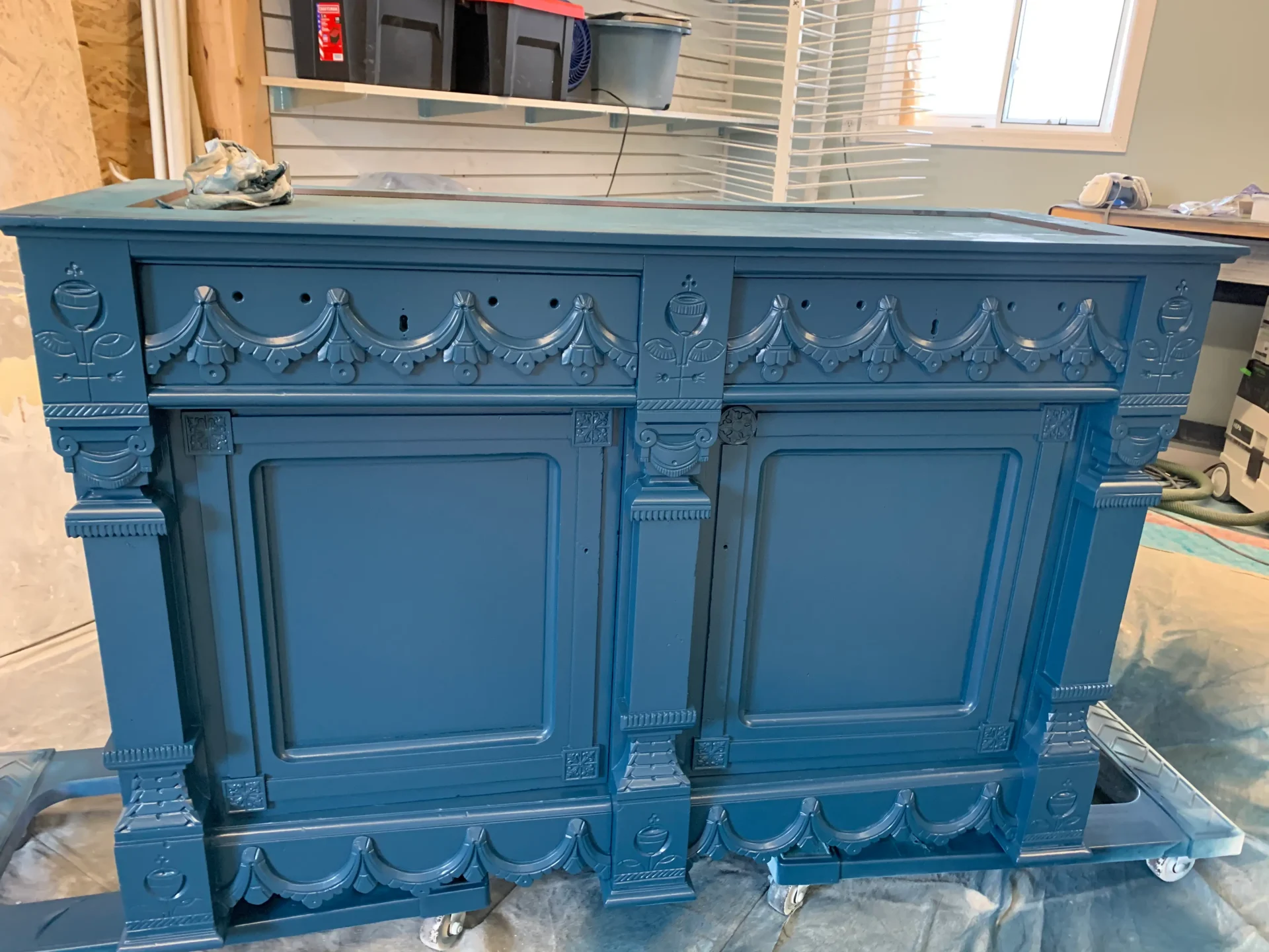 A blue, ornately carved wooden cabinet sits on a plastic-covered floor in a workshop, with paint supplies nearby and shelves in the background. - Jeff Bryant Painting