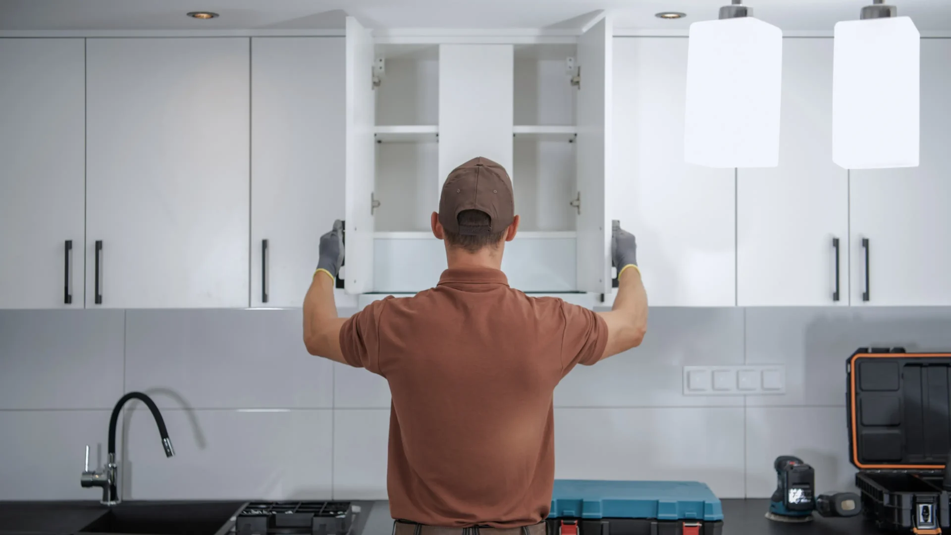A person wearing gloves and a cap opens the doors of an empty kitchen cabinet, with tools and equipment on the countertop below. - Jeff Bryant Painting