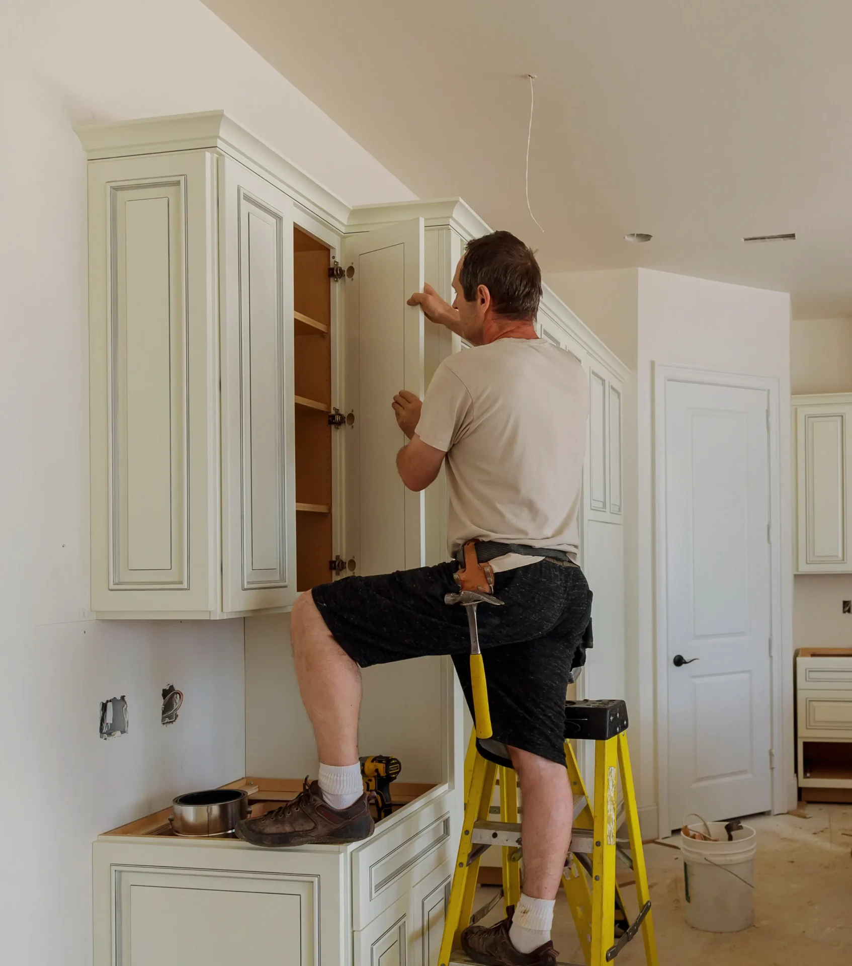 A man stands on a yellow ladder installing or adjusting a cabinet door in a kitchen under renovation. - Jeff Bryant Painting