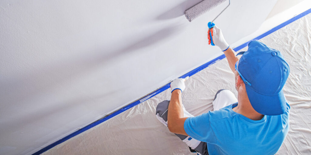A person in blue clothing and gloves uses a paint roller to paint a white wall, with a drop cloth and painter's tape protecting the floor and trim. 