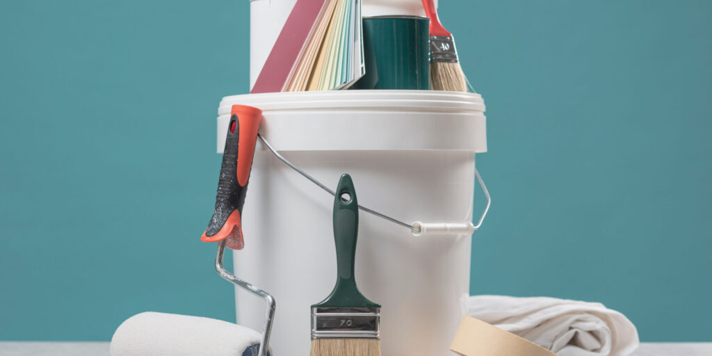 White paint bucket with painting tools like paintbrushes, a roller, paint swatches, a sanding block, sandpaper, and a cloth set against a teal background. - Jeff Bryant Painting