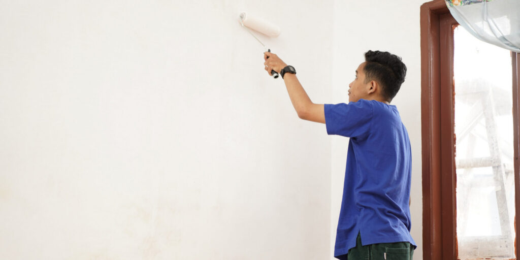 A person in a blue shirt uses a paint roller to paint a white wall near a window, with natural light brightening the room. - Jeff Bryant Painting