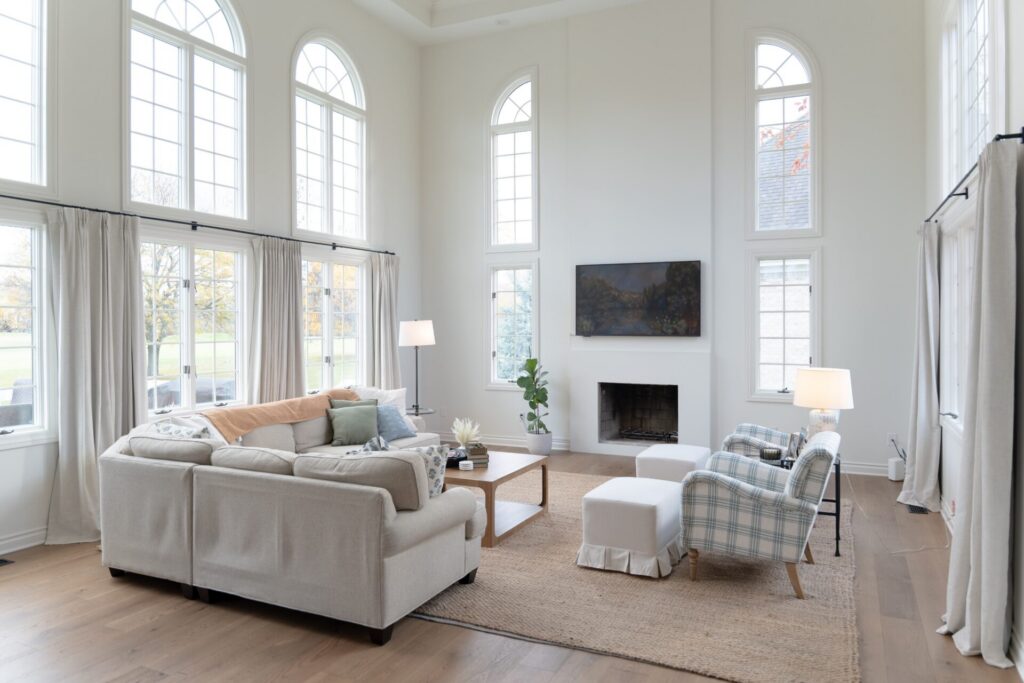 A spacious living room with tall arched windows, light-colored furniture, a fireplace, two lamps, and a wall-mounted TV. Recently refreshed by expert interior painting in Alto Michigan, natural light beautifully fills the space. - Jeff Bryant Painting