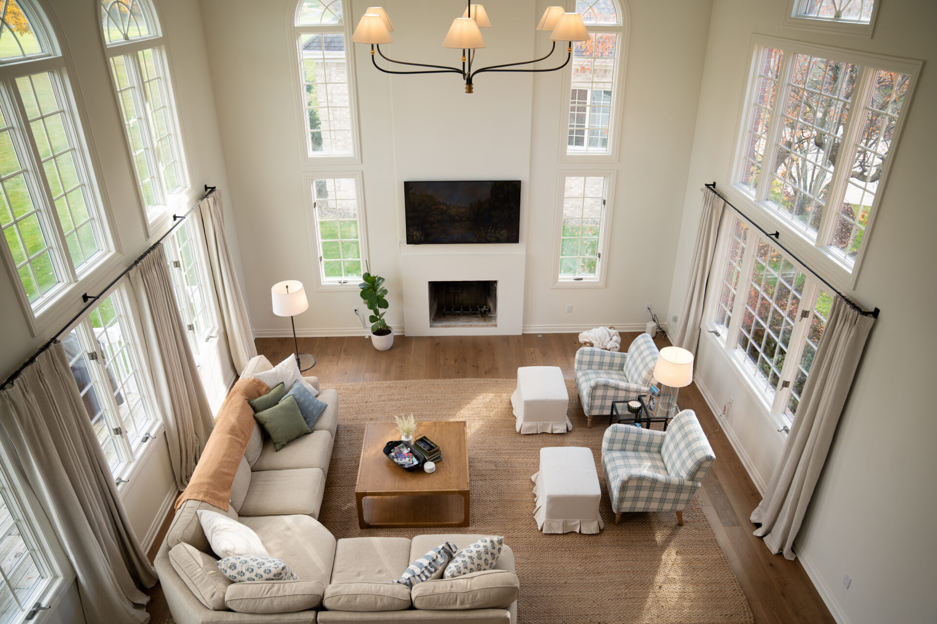 A spacious living room with large windows, beige sectional sofa, two checkered armchairs, and a fireplace with a TV above it—showcasing the fresh look from recent interior painting in Alto, Michigan—viewed from above. - Jeff Bryant Painting
