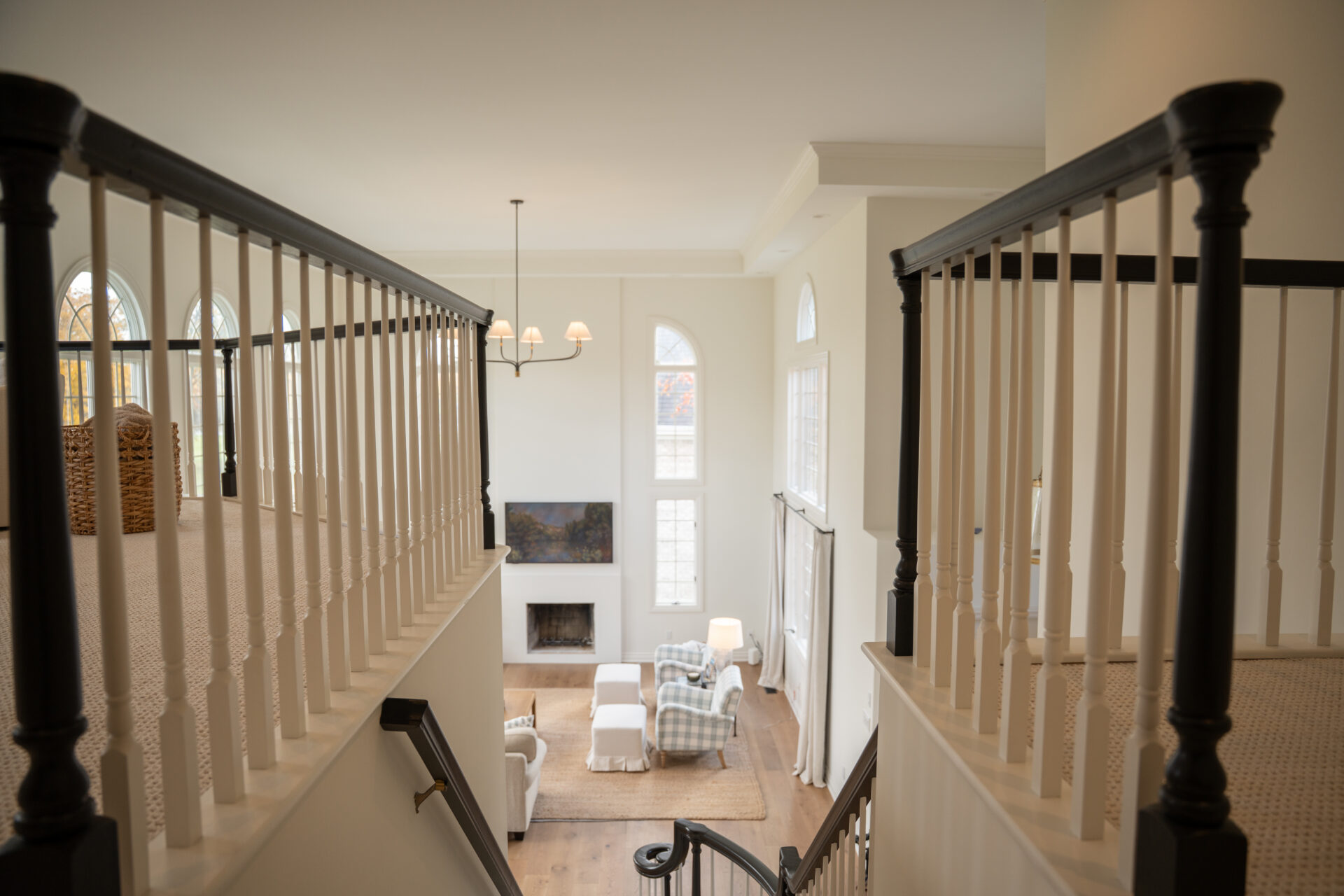 View from staircase landing overlooking a living room with tall windows, a fireplace, armchairs, TV, and light neutral decor—showcasing expert interior painting in Alto Michigan. - Jeff Bryant Painting