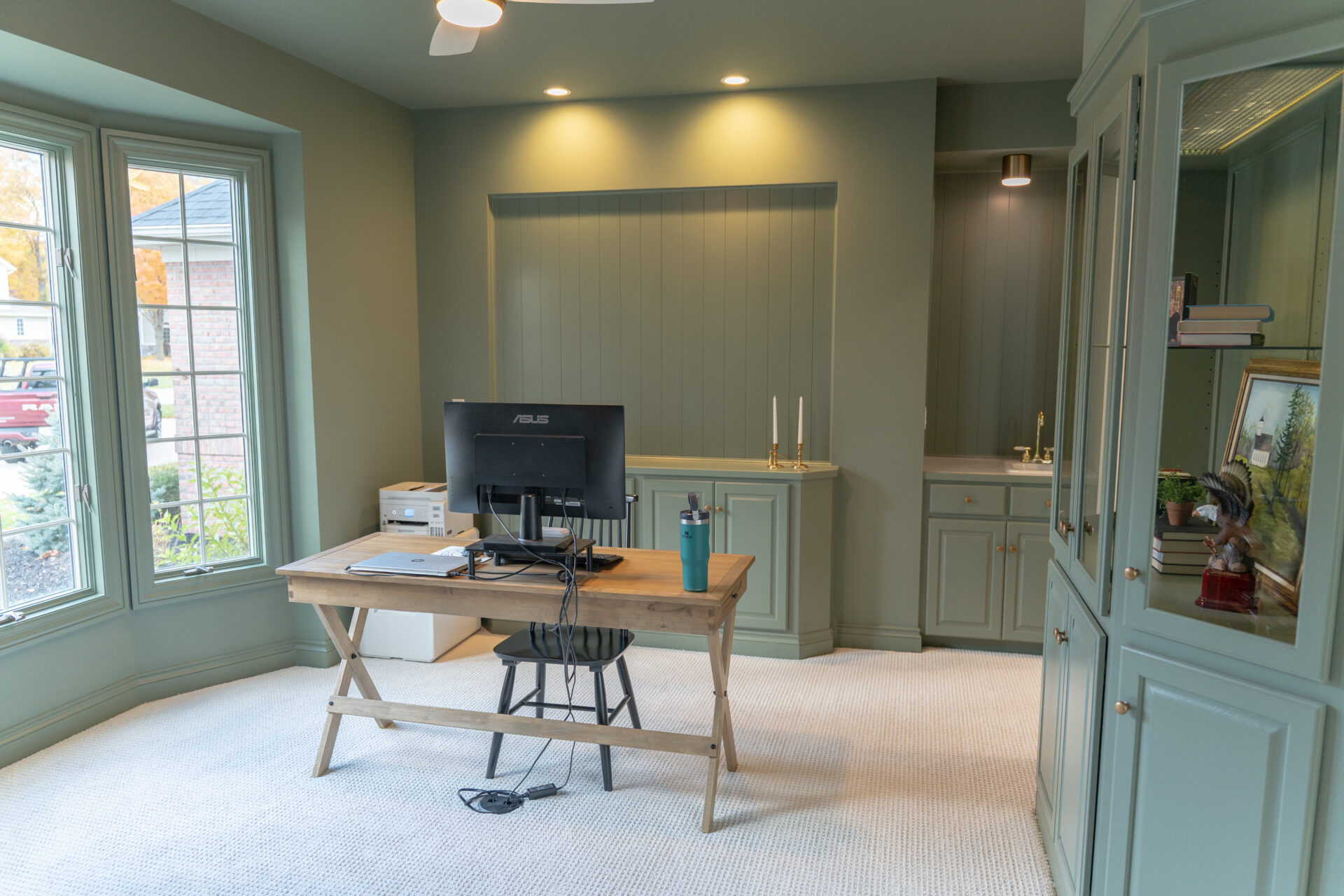 A home office with green cabinetry from an interior painting Alto Michigan project, featuring a wooden desk with a computer monitor, chair, water bottle, large windows, and built-in shelves. - Jeff Bryant Painting